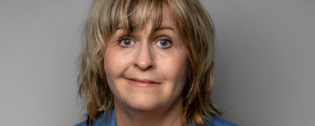 Person med axellångt blont hår, blå kavaj och vit topp mot grå bakgrund.