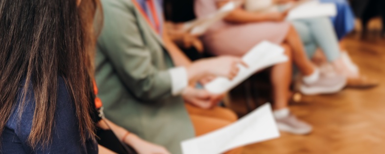 En grupp människor sitter i rad på stolar och håller papper, troligen under ett möte eller en workshop.