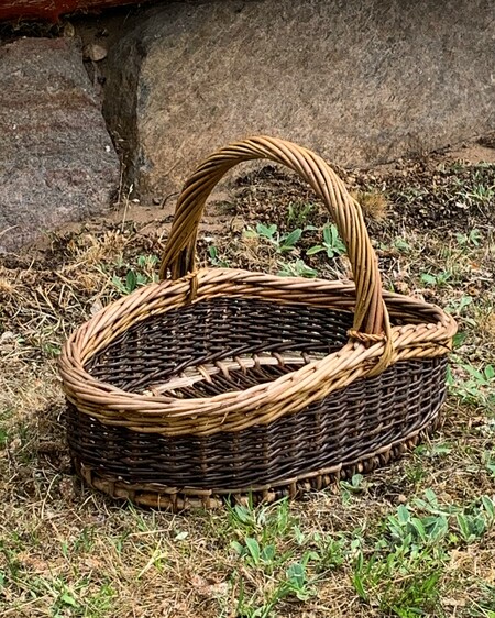 En tom, oval korg av flätad pil står på gräs vid en stenmur utomhus.