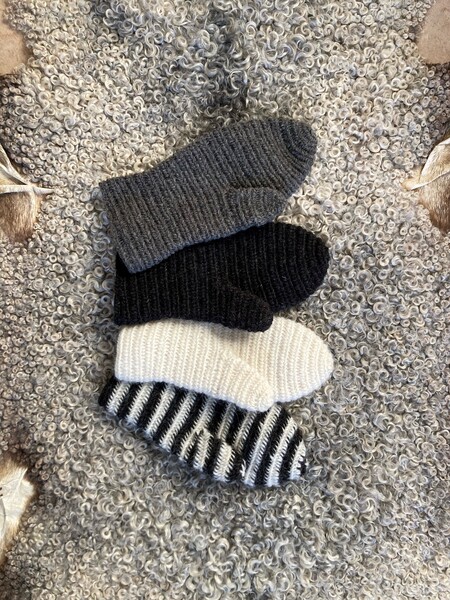 Fyra nålbundna vantar i grått, svart, vitt och svartvitt randigt ligger på en lockig grå ullpläd.