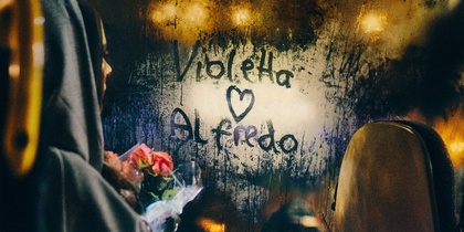 En person med huva och blombukett sitter på en buss, med texten 'Violetta ♥ Alfredo' skriven på en immig fönsterruta.