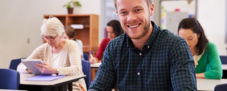 Leende man sitter vid skolbänk i klassrum, skriver i anteckningsbok. Andra vuxna studenter i bakgrunden.
