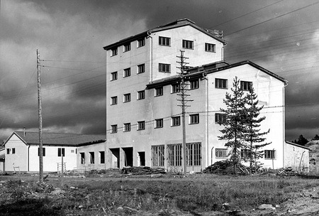 En svartvit bild på ett stort vitt hus med fyra våningar. En mörkt molning himmel i bakgrunden. Framför huset telefonstolpar med kablar. 