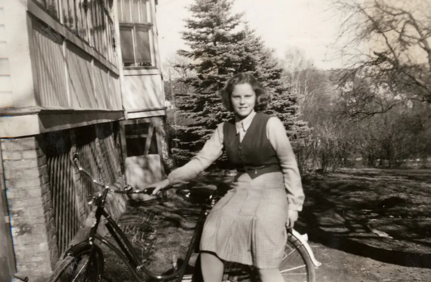 Kvinna sitter på en cykel utomhus bredvid ett hus, med träd i bakgrunden, i en svartvit bild.