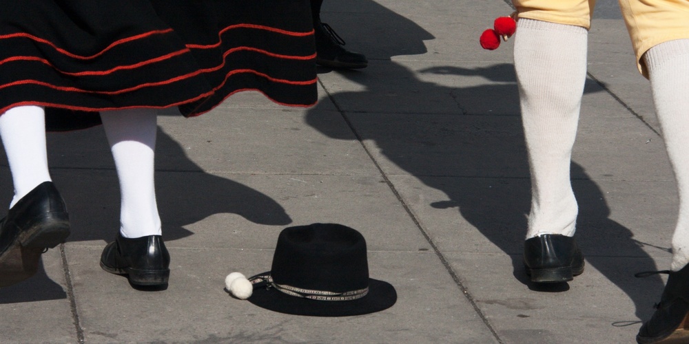 Två personer i traditionella folkdräkter dansar på en stenlagd yta, en svart hatt ligger på marken.