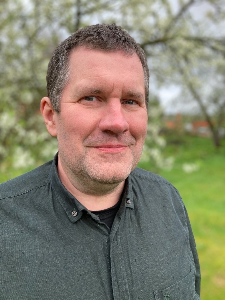 Lars-Gunnar Jonsson med mörkgrön skjorta står utomhus framför blommande träd.