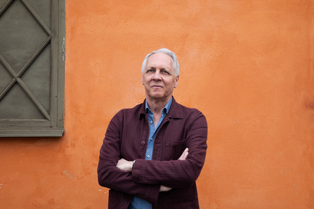 Äldre man med grått hår står med armarna i kors framför en orange vägg bredvid ett fönster.