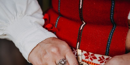 Närbild av händer som håller en traditionell väska med röd brodyr och mässingsdetaljer, klädd i folkdräkt.