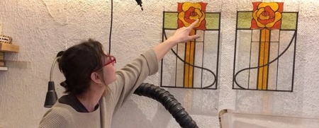 En kvinna pekar på ett färgglatt glasfönster med rosmotiv i en verkstadsmiljö.