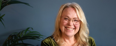 Leende äldre kvinna med långt blont hår och glasögon sitter mot blå vägg, grön tröja och växt i bakgrunden.