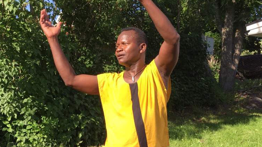 Man i gul och svart tröja står utomhus med armarna uppsträckta mot himlen.