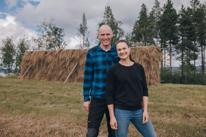 Björn Ferry och Heidi Andersson står på en äng framför en stor hög med hö, omgivna av träd och molnig himmel.