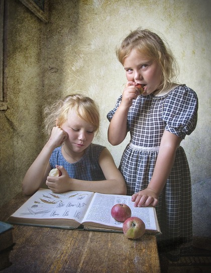 Två flickor i rutiga klänningar sitter vid ett bord med en bok och äpplen, en flicka äter ett äpple.