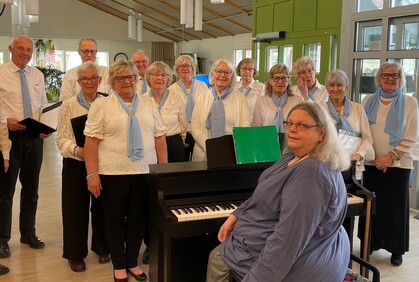 En grupp äldre personer står samlade runt ett piano i en ljus lokal, klädda i vitt och blått.