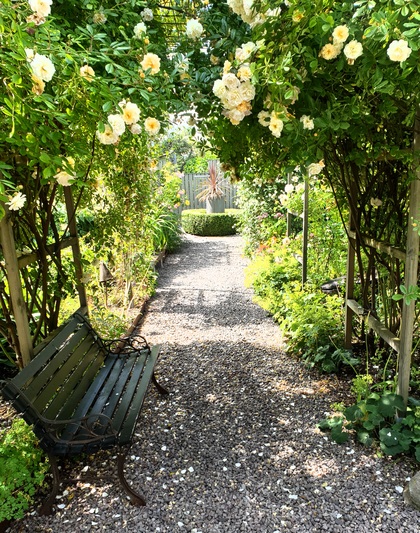 En grusgång i en blomstrande trädgård med en bänk under en rosbåge.