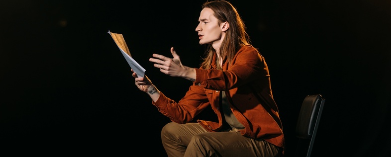 En person sitter på en stol på en scen och gestikulerar med ett manus i handen under en teaterövning.