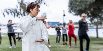 En grupp människor utövar tai chi utomhus i en park, med en kvinna i vit dräkt i förgrunden.