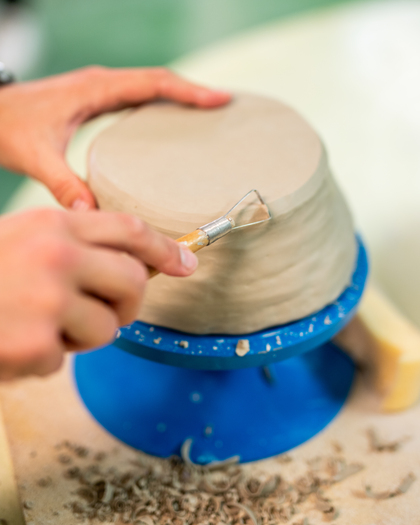 En person beskär en lerkruka med ett verktyg på en blå drejskiva.