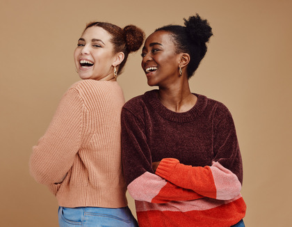 Två unga kvinnor står rygg mot rygg och skrattar mot kameran, mot beige bakgrund.