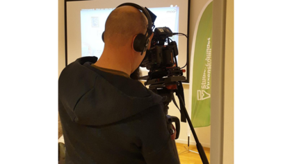 En person med hörlurar filmar med en videokamera i ett inomhusmiljö, framför en projektorduk.