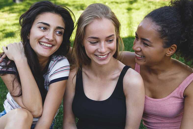 Tre leende tonåringar sitter tätt tillsammans utomhus på gräset.