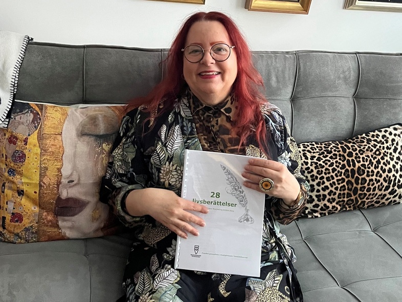 Kvinna med rött hår och glasögon sitter på en soffa och håller ett dokument med texten '28 läsberättelser'.