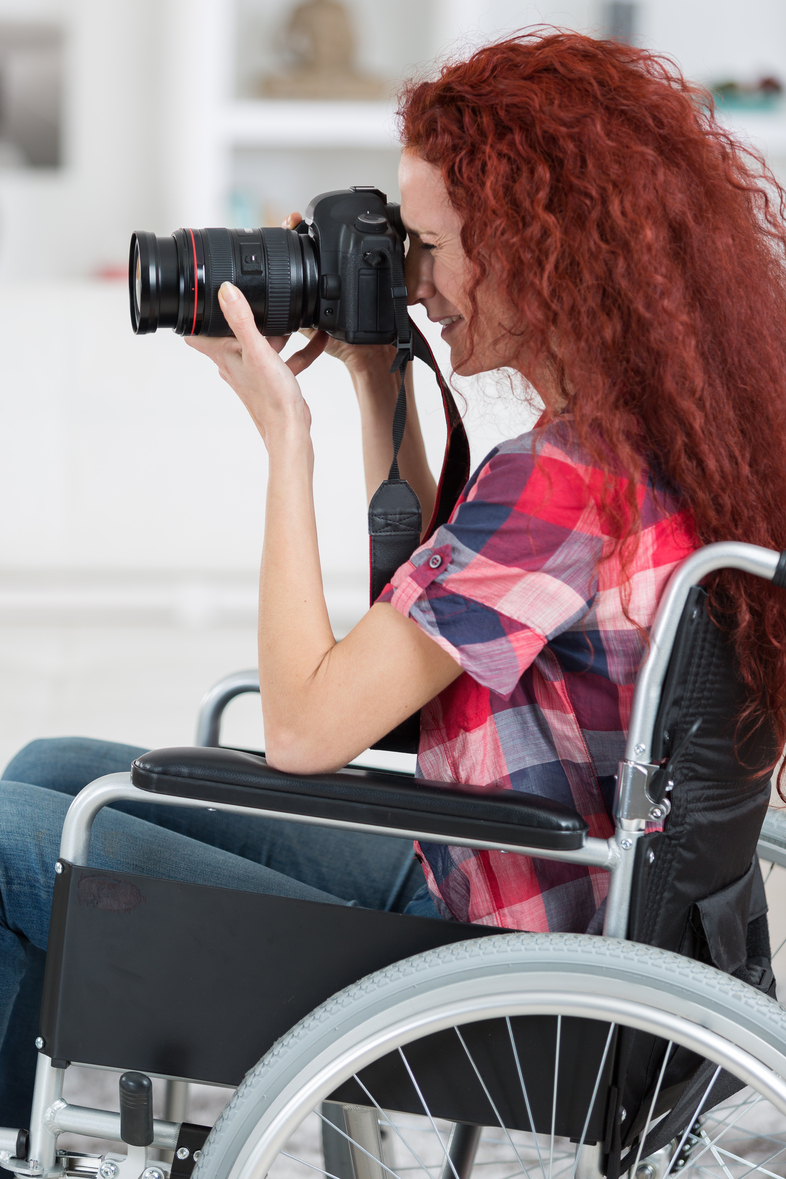 En person med långt rött hår sitter i rullstol och fotograferar med en systemkamera.
