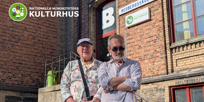 Två äldre män står utanför en tegelbyggnad med skyltar för Redbergsskolan och Nationella minoriteters kulturhus.