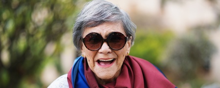 Leende äldre kvinna med grått hår, stora solglasögon och färgglad sjal utomhus.