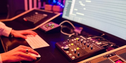 En person arbetar vid en dator i en musikstudio med ljudutrustning och mixerbord.