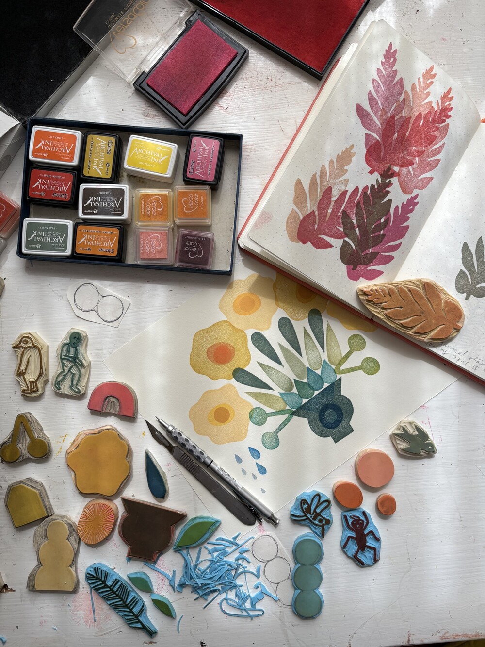 Ett skrivbord med stämplar, färgkuddar, verktyg och en öppen skissbok med bladmotiv.