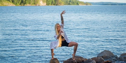 En kvinna med långt blont hår poserar i en dansställning på stenar vid en sjö, omgiven av vatten och skog.