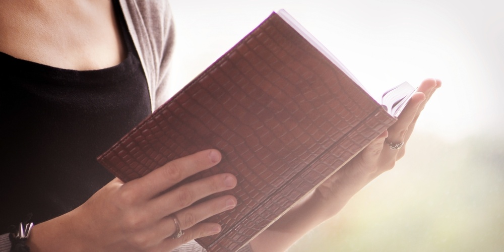 En person håller och läser en bok med brun pärm, endast överkroppen och händerna syns.