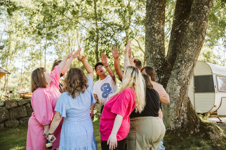 En grupp människor står i en ring utomhus och sträcker upp händerna mot mitten under ett träd.