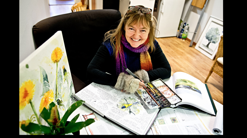 Person målar i en bok med akvarellfärger, omgiven av konst och böcker i ett ljust rum.