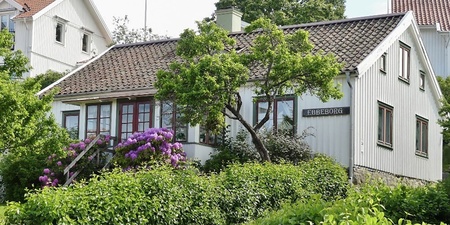 Vit trävilla med blommande buskar och trädgård, sommargrönska och grusgång framför huset.