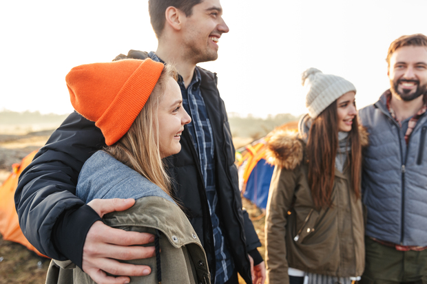 Fyra unga vuxna står tillsammans utomhus vid tält, klädda i varma kläder och mössor, och ser glada ut.