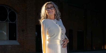 Kvinna i ljus kjol och tröja står på en perrong med tegelvägg och stora fönster i bakgrunden.