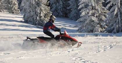 En person kör en röd snöskoter på ett snötäckt fält med frostiga granar i bakgrunden.