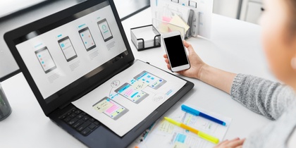 Person arbetar med appdesign på laptop och smartphone vid ett skrivbord med anteckningar och färgpennor.