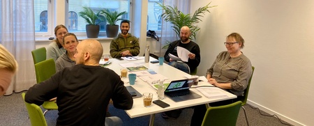 Sex personer sitter runt ett konferensbord i ett ljust kontorsrum med stora fönster och gröna växter.