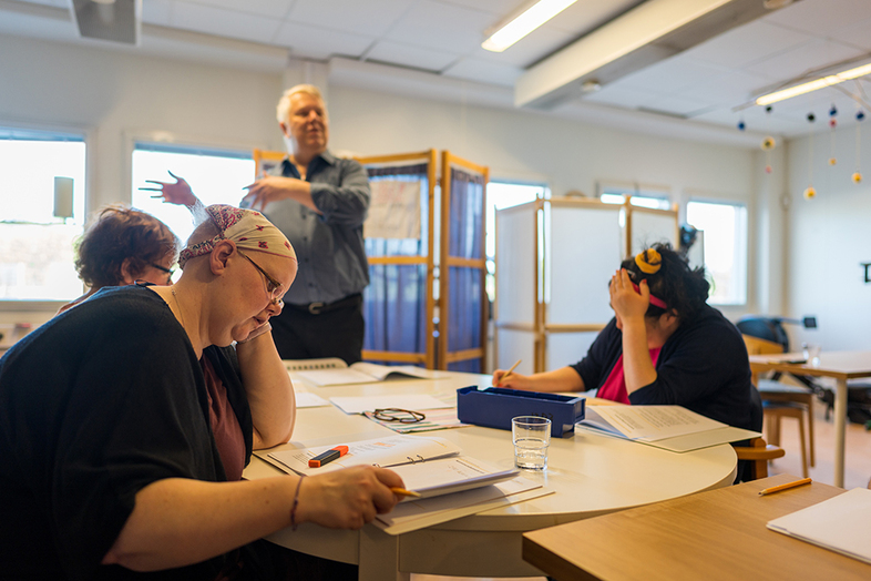 Tre personer sitter vid ett bord och arbetar med papper, en person står och gestikulerar i bakgrunden.