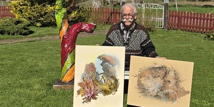 Person sitter utomhus och visar två konstverk, med en färgglad träskulptur bredvid.