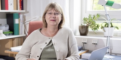 Person sitter vid ett skrivbord med laptop, böcker och växter i ett kontorsrum.