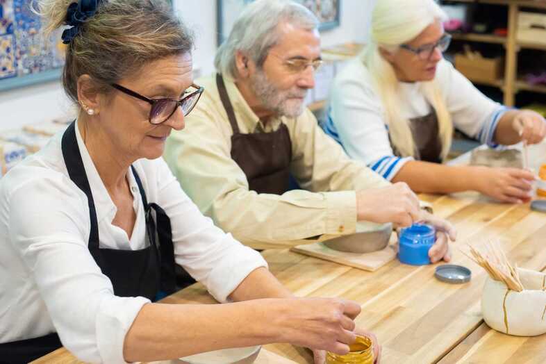 Tre äldre personer har på sig förkläden och sitter vid ett bord och pysslar.