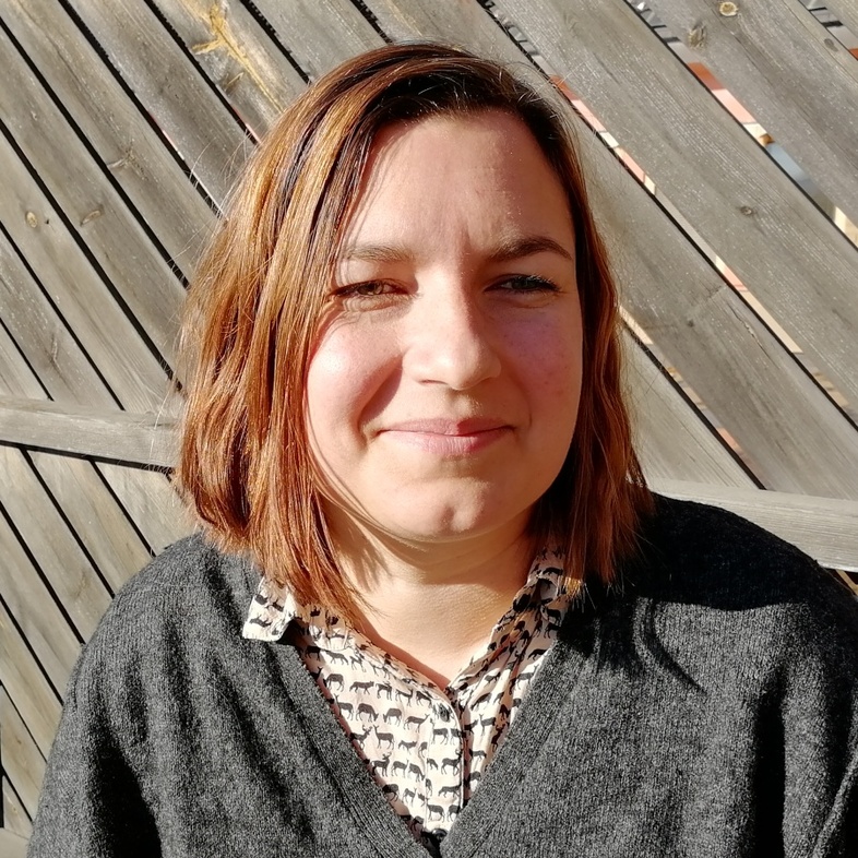 Kvinna med axellångt hår sitter utomhus i solen framför ett trästaket.