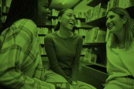 Tre unga kvinnor sitter och skrattar tillsammans i ett bibliotek.