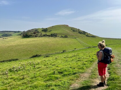 En person med ryggsäck står på en stig och tittar ut över gröna kullar under en klar himmel.