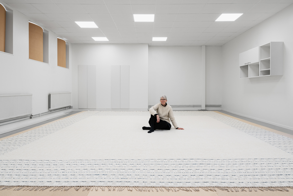 En person sitter på ett stort, ljust matta i ett minimalistiskt, vitt rum med högt i tak.