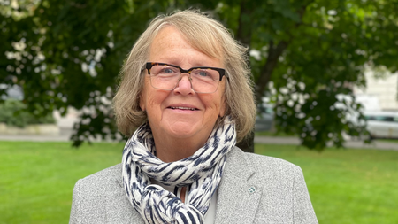 Äldre kvinna med glasögon och grå kavaj står utomhus framför gröna träd och gräsmatta.